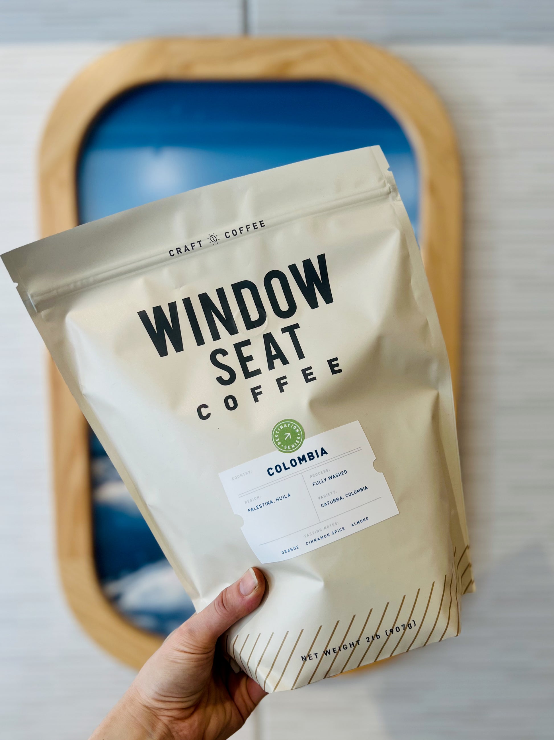 Two pound bag of single-origin Colombia coffee from the Huila region held in front of a window seat painting. Fully washed processed with tasting notes of orange, cinnamon spice, and almond.