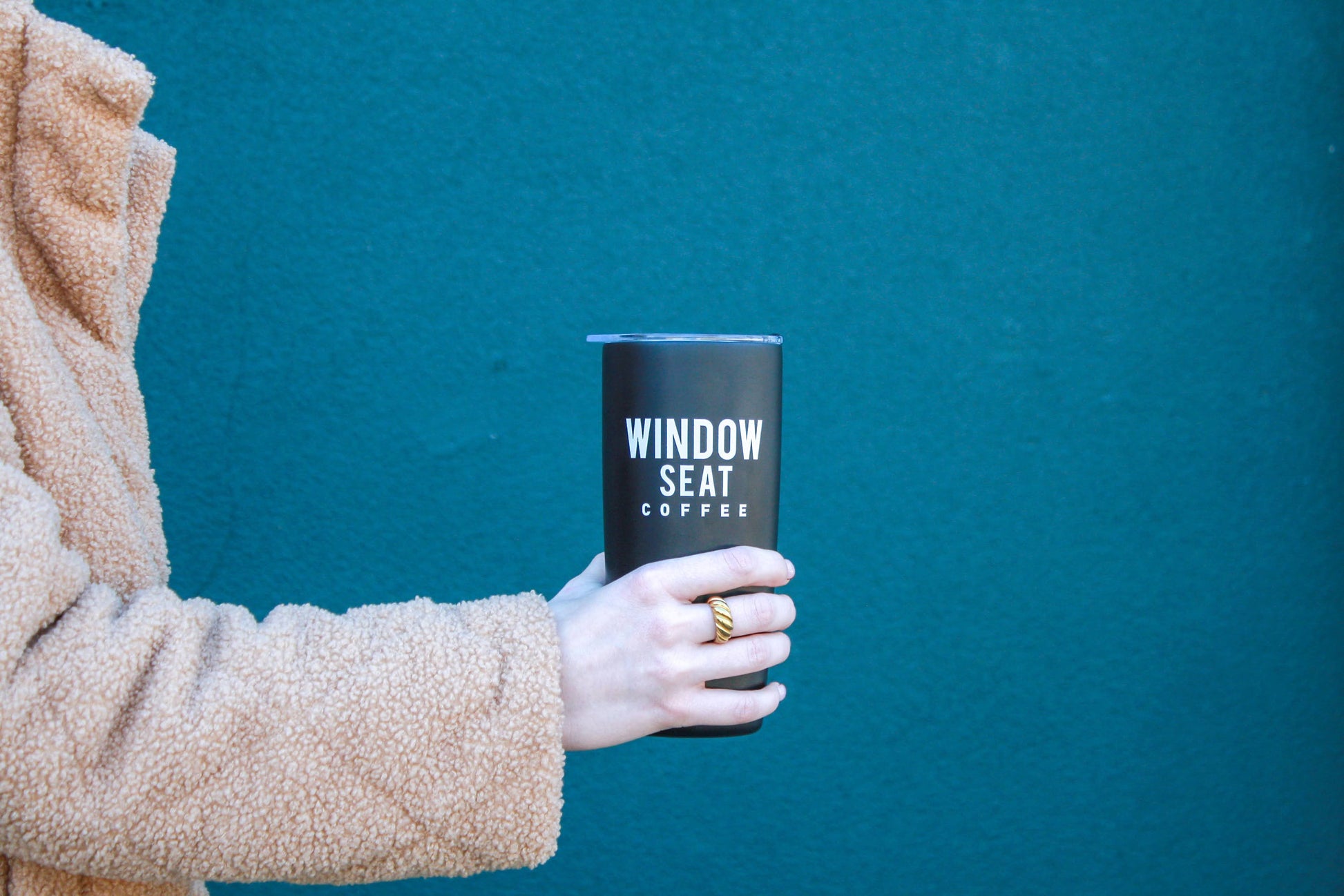 Hand holding Black 20oz travel tumbler by Window Seat Coffee and MIIR against green wall